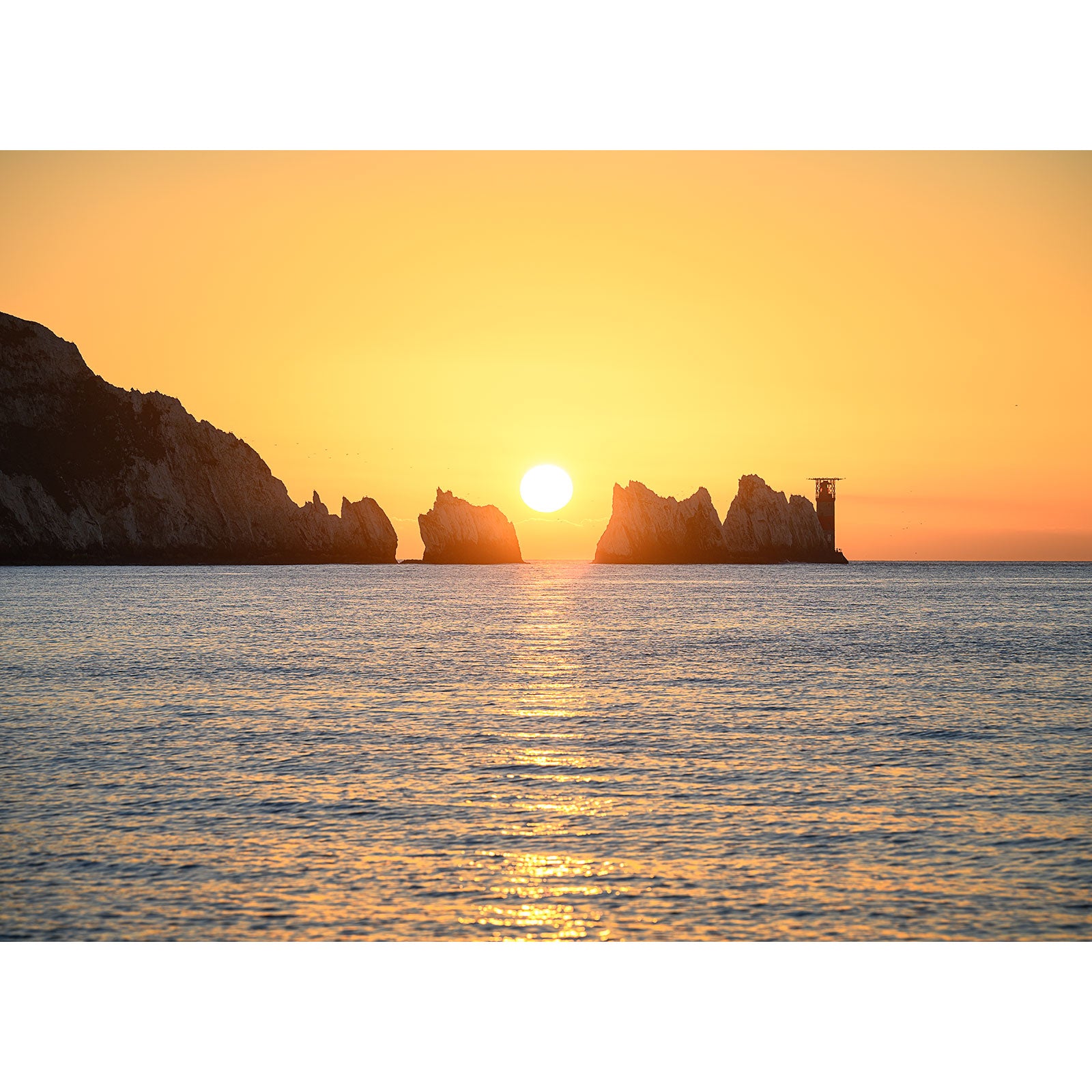 Sunset over the ocean with the sun positioned between rock formations and a silhouette of a lookout tower on the right, near The Needles by Available Light Photography.