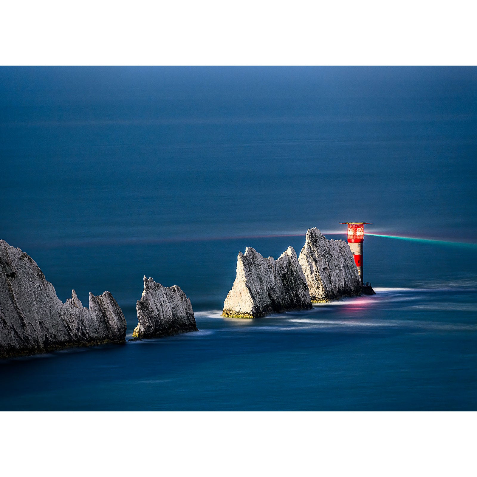 The Needles by Moonlight lighthouse stands at the end of a series of jagged white cliffs extending into a blue sea on the Isle of Wight, captured beautifully by Available Light Photography.