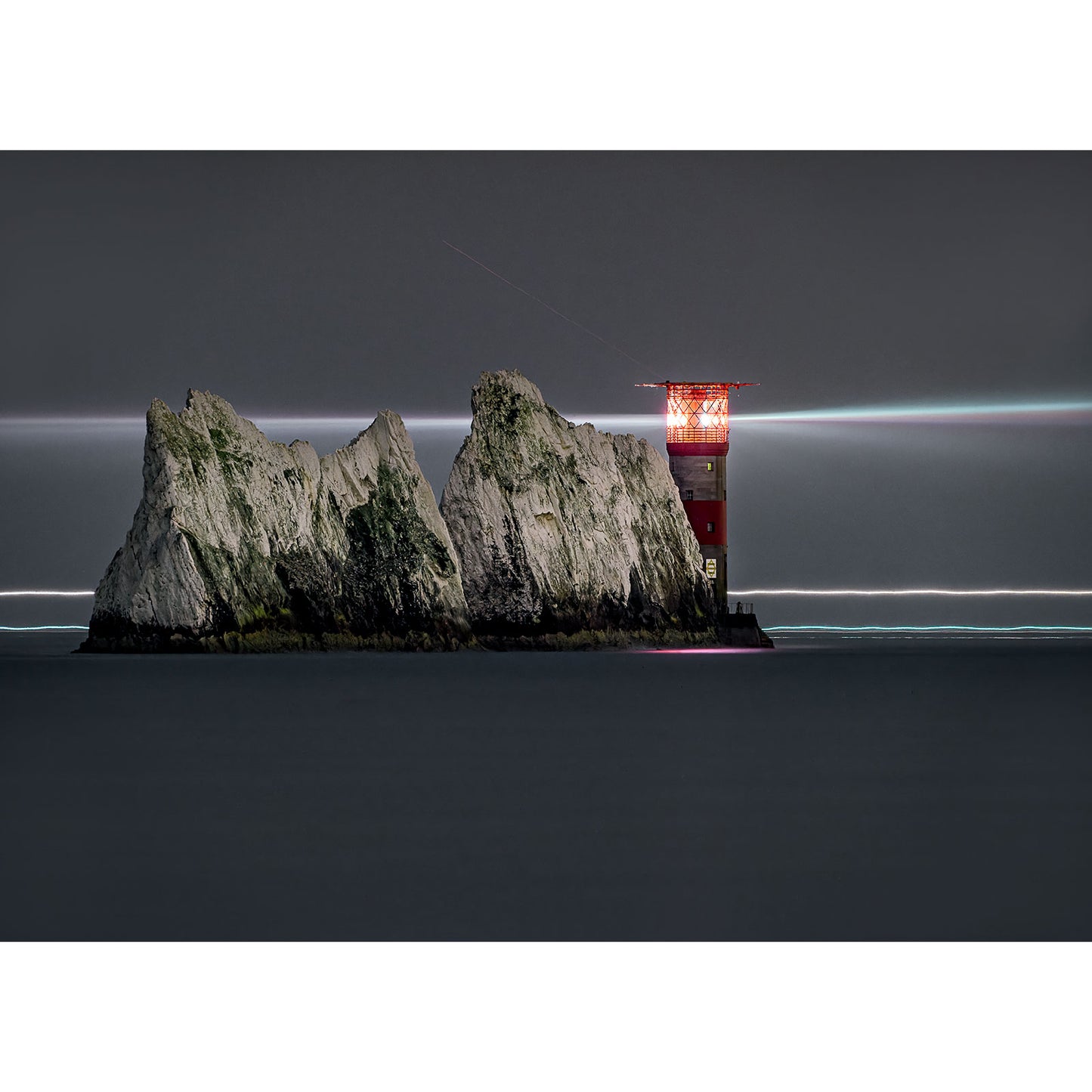A lighthouse atop a craggy isle at night, with a long exposure capturing a streak of light from a passing ship by Available Light Photography's The Needles.