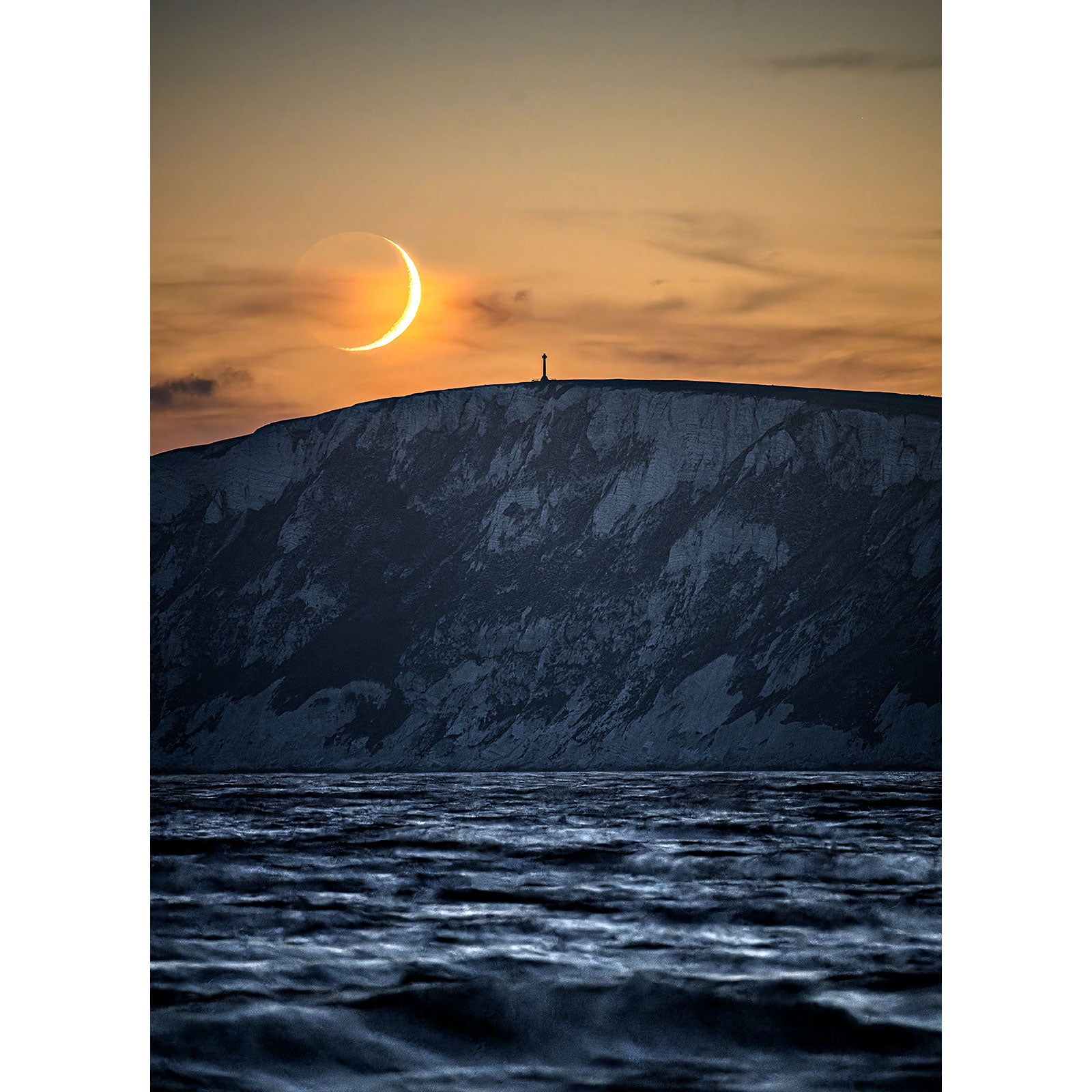 A person stands at the edge of a snow-covered cliff on the Isle of Wight with a Crescent Moon occurring behind them.