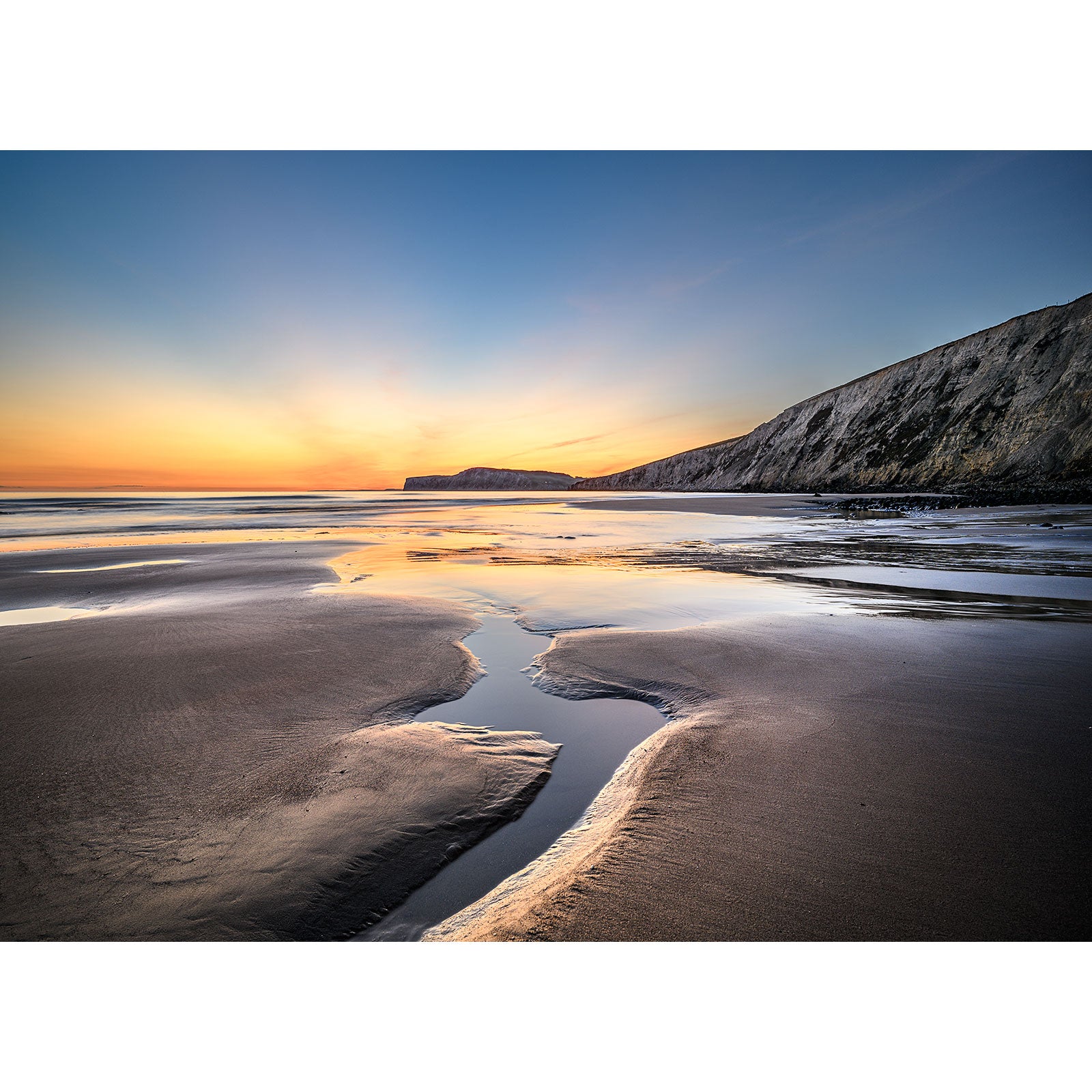 A serene sunset at a coastal beach with water channels in the sand and cliffs in the background, reminiscent of a scene captured by Available Light Photography's Compton Bay.