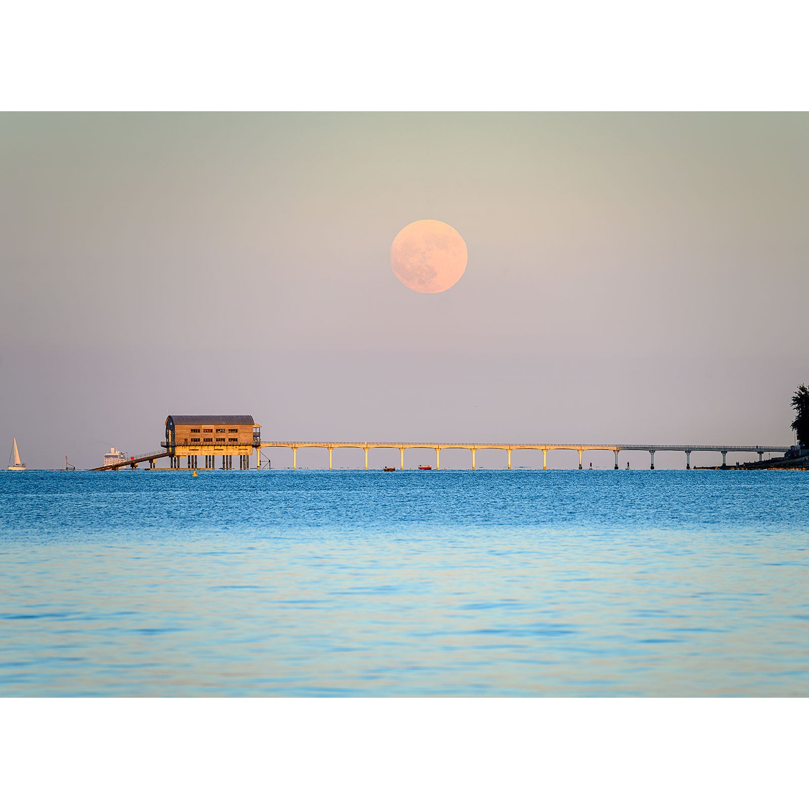 A Moonrise over Bembridge Lifeboat Station rises over a tranquil sea with a pier in the background during twilight on the Isle of Wight, captured beautifully by Available Light Photography.