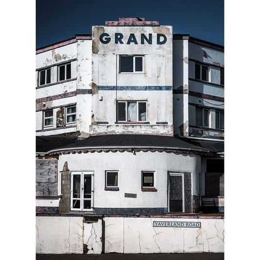 A dilapidated building labeled "The Grand, Sandown" on a sunny day, showcasing signs of neglect and decay, at the intersection of a road named "tavernland" on the Isle of Wight. By Available Light Photography.