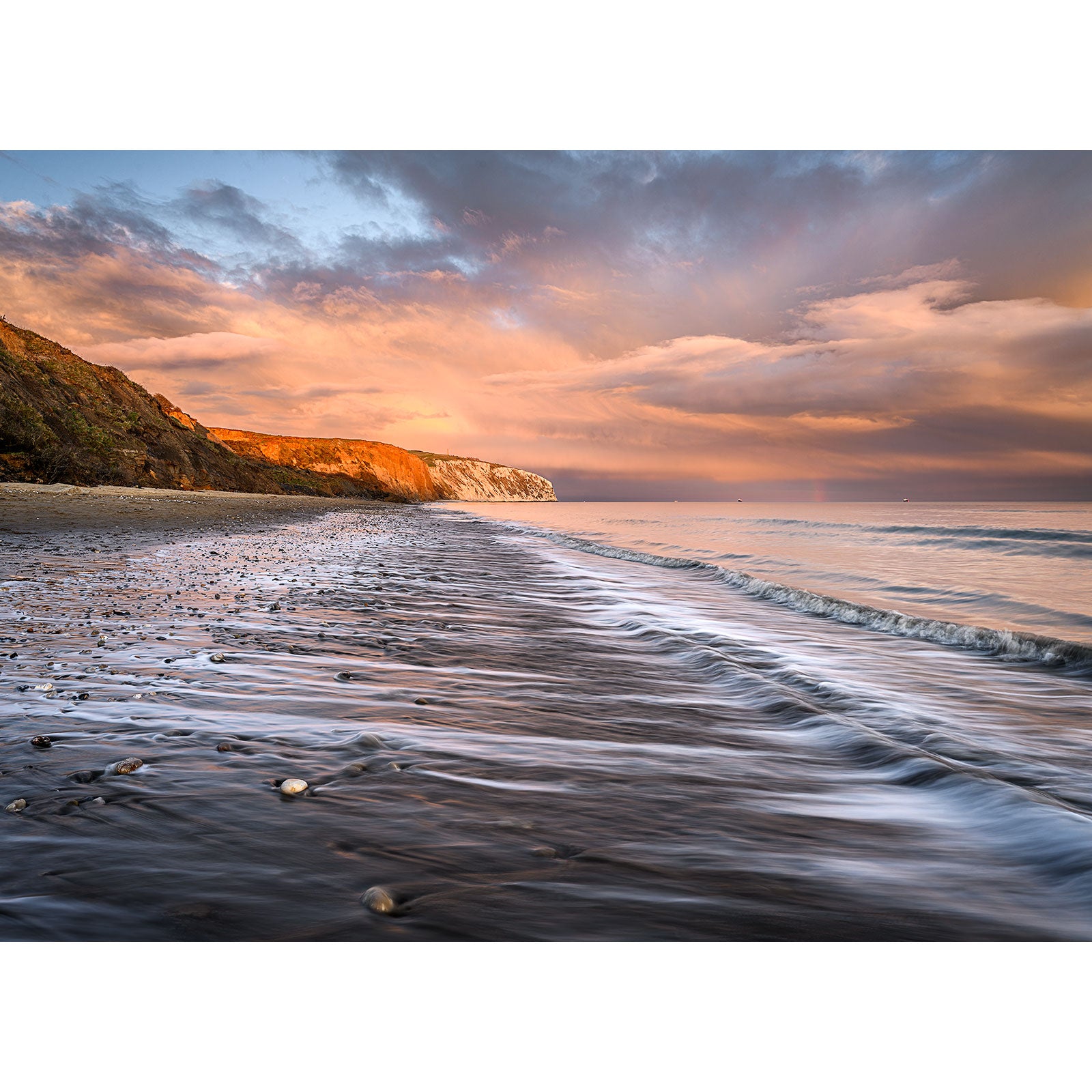 Sunset paints the sky with warm hues over a serene beach on the Isle of Wight, highlighting the contours of Culver Cliff and the gentle wash of waves on the sand. (Product Name: Culver Cliff, Brand Name: Available Light Photography)