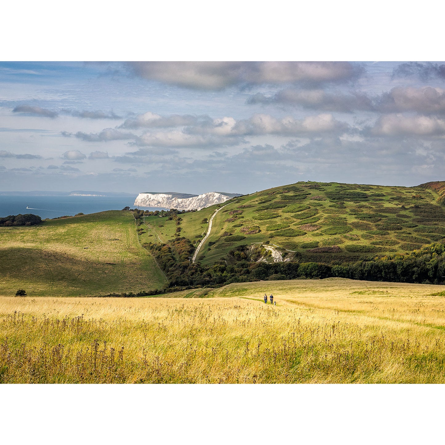Two people walking on a path through rolling grassy hills with cliffs in the distance on the Isle of Gascoigne, captured in "View over West Wight" by Available Light Photography.