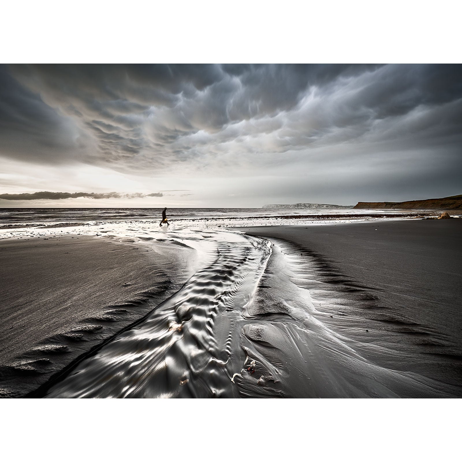 A solitary figure walks along Brook Bay beach on the Isle of Wight with rippling waters under a dramatic cloudy sky captured by Available Light Photography.