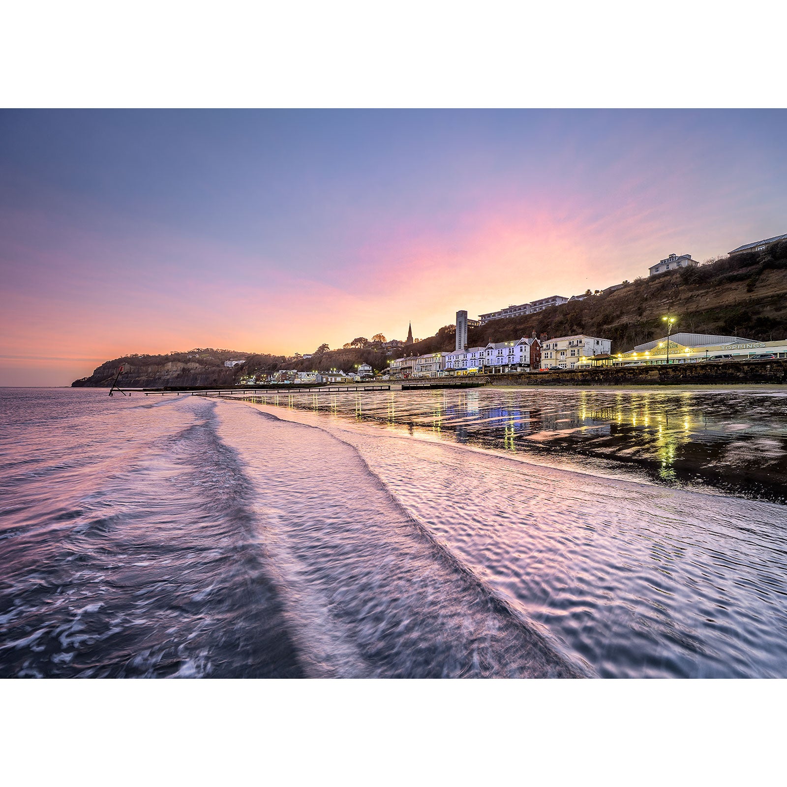 Coastal town at dusk with lit buildings reflecting on wet sand under a pastel-colored sky on Shanklin Beach by Available Light Photography.