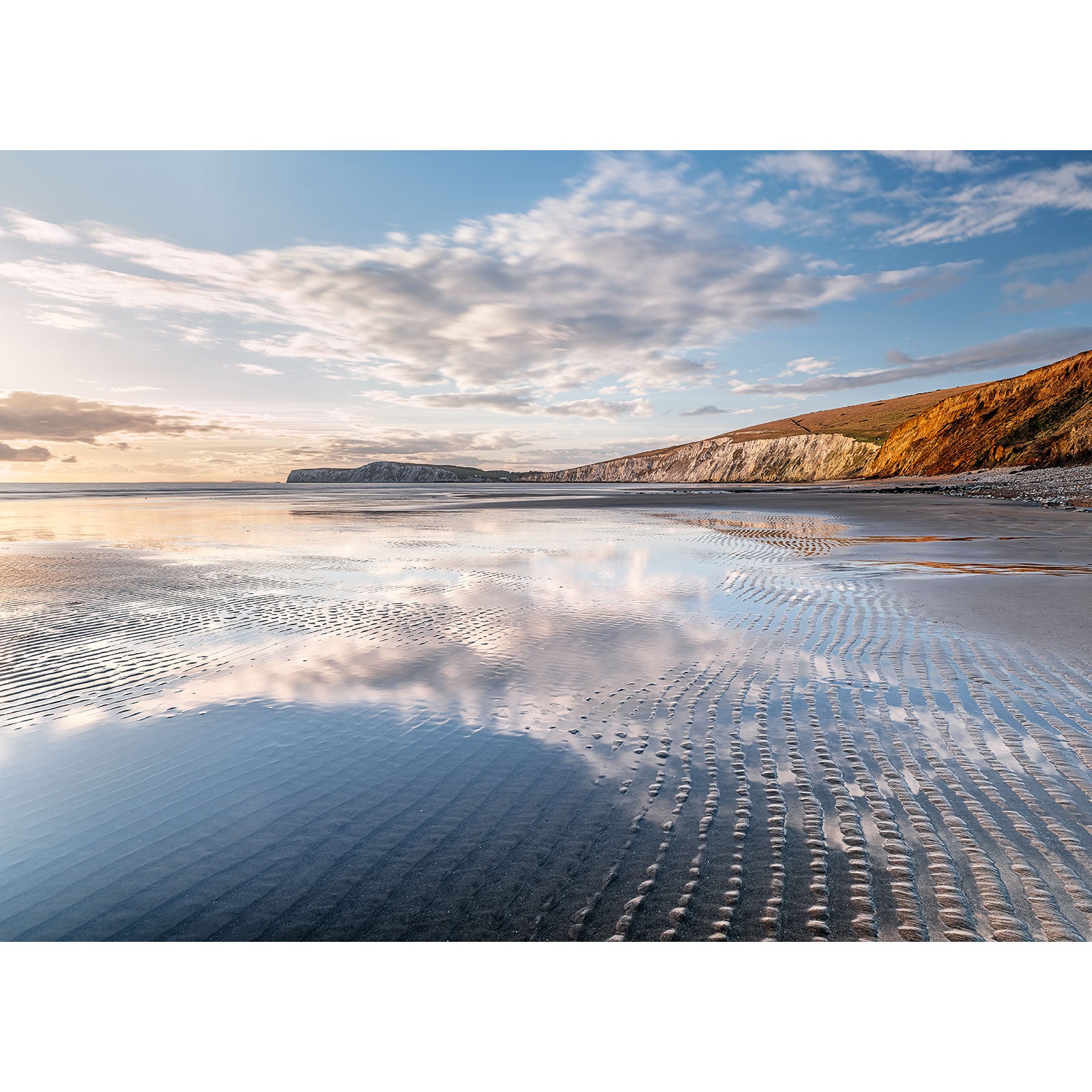 A serene beach at low tide with rippled sand, coastal cliffs in the background, and a calm sunset sky over Compton Bay by Available Light Photography.