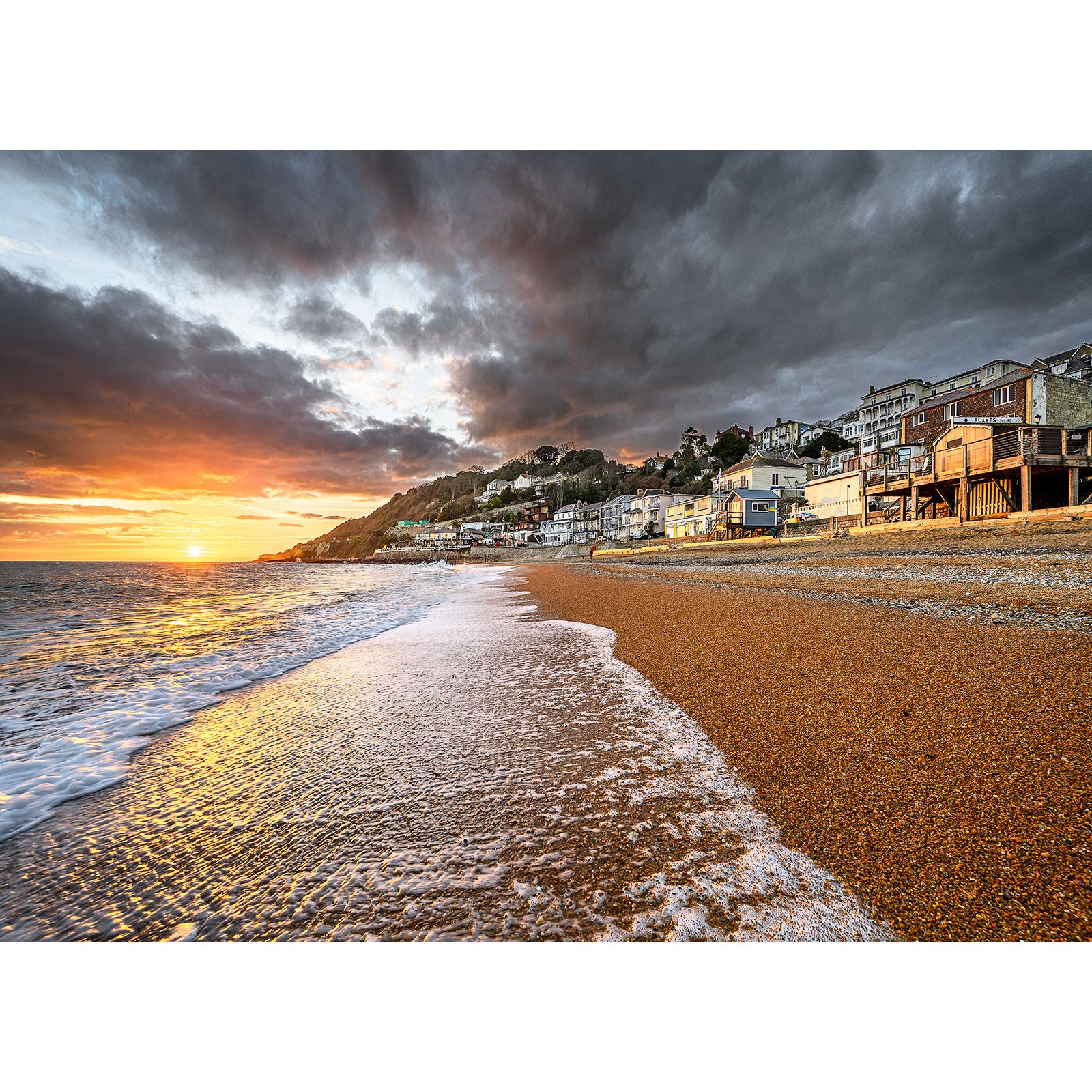 Sunset at a pebble beach on the Isle of Ventnor with a coastal town in the background and dramatic clouds overhead, captured by Available Light Photography.