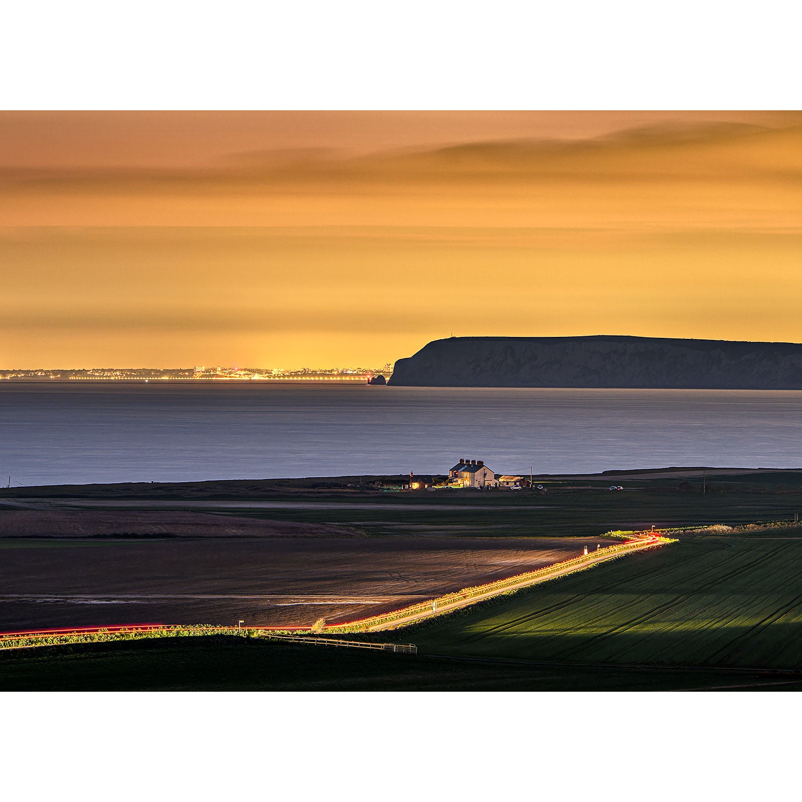 A serene landscape at dusk on the Isle of Wight with layers of farmland, a small building in the foreground, and a cliff in the distance under a gradient sunset sky captured by Looking toward The Needles by Available Light Photography.