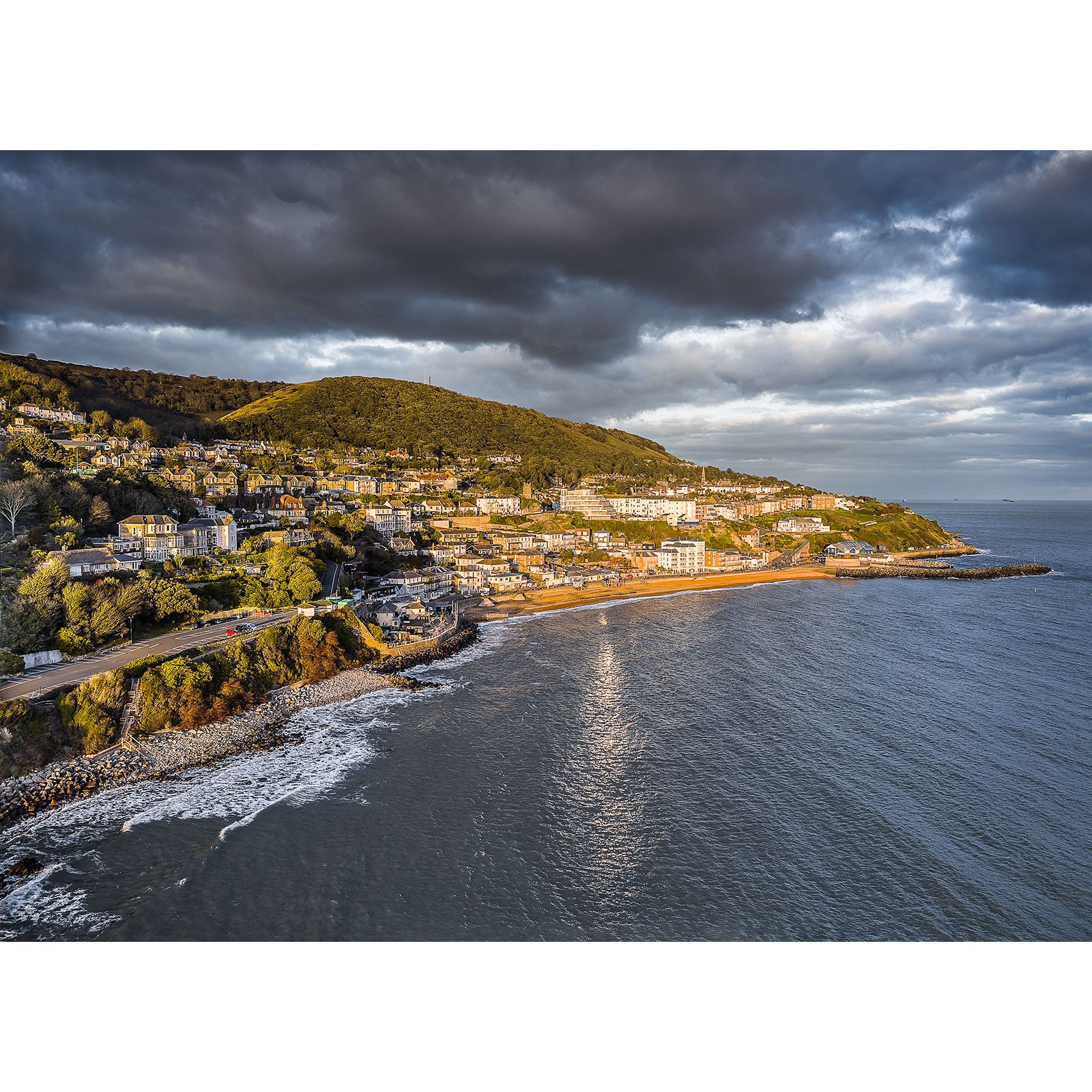 Coastal town bathed in sunlight with a dramatic cloud-covered sky overhead on the Isle of Ventnor by Available Light Photography.