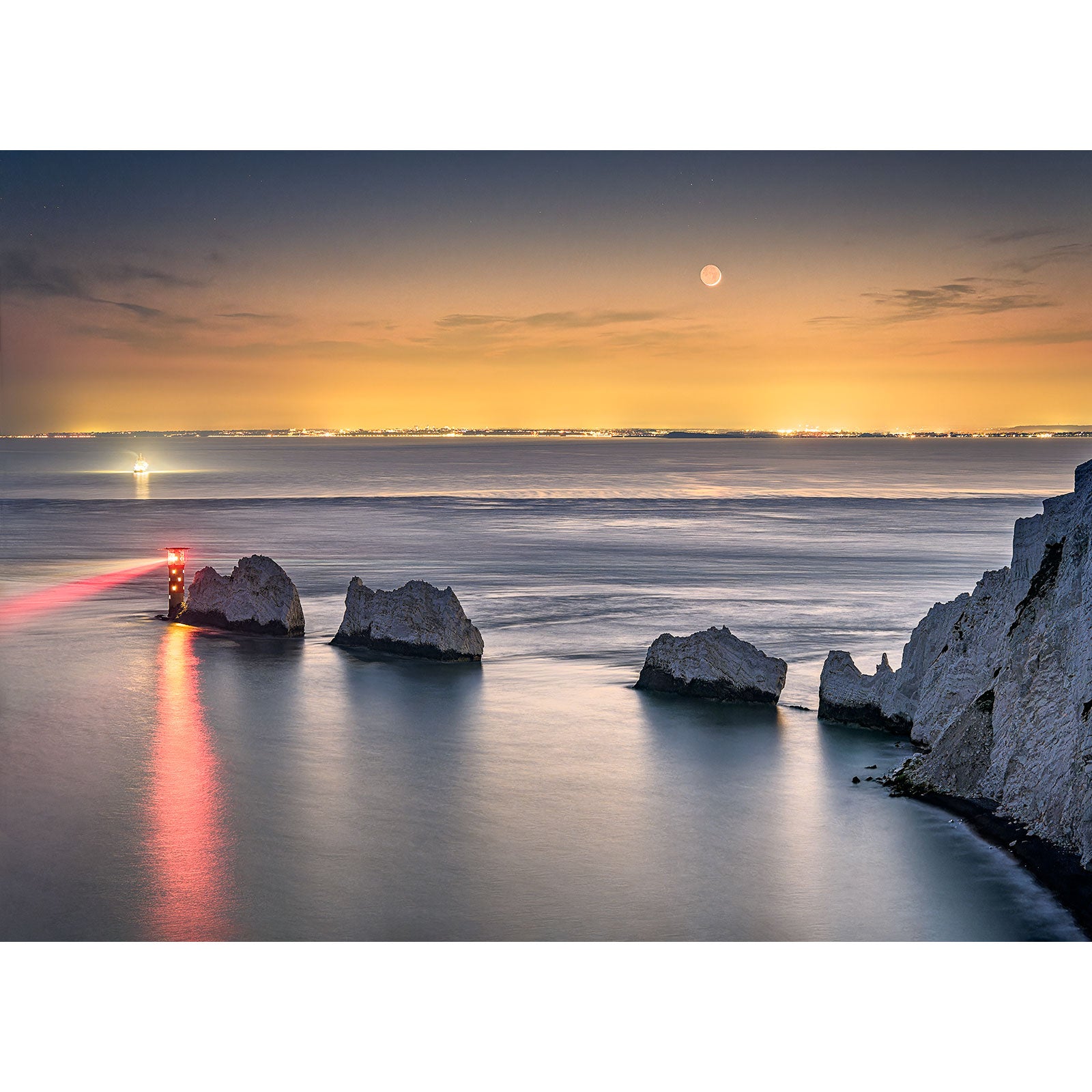 Seascape at twilight with a lighthouse beam and Moonset over The Needles and moon over calm waters and chalk cliffs of Wight by Available Light Photography.
